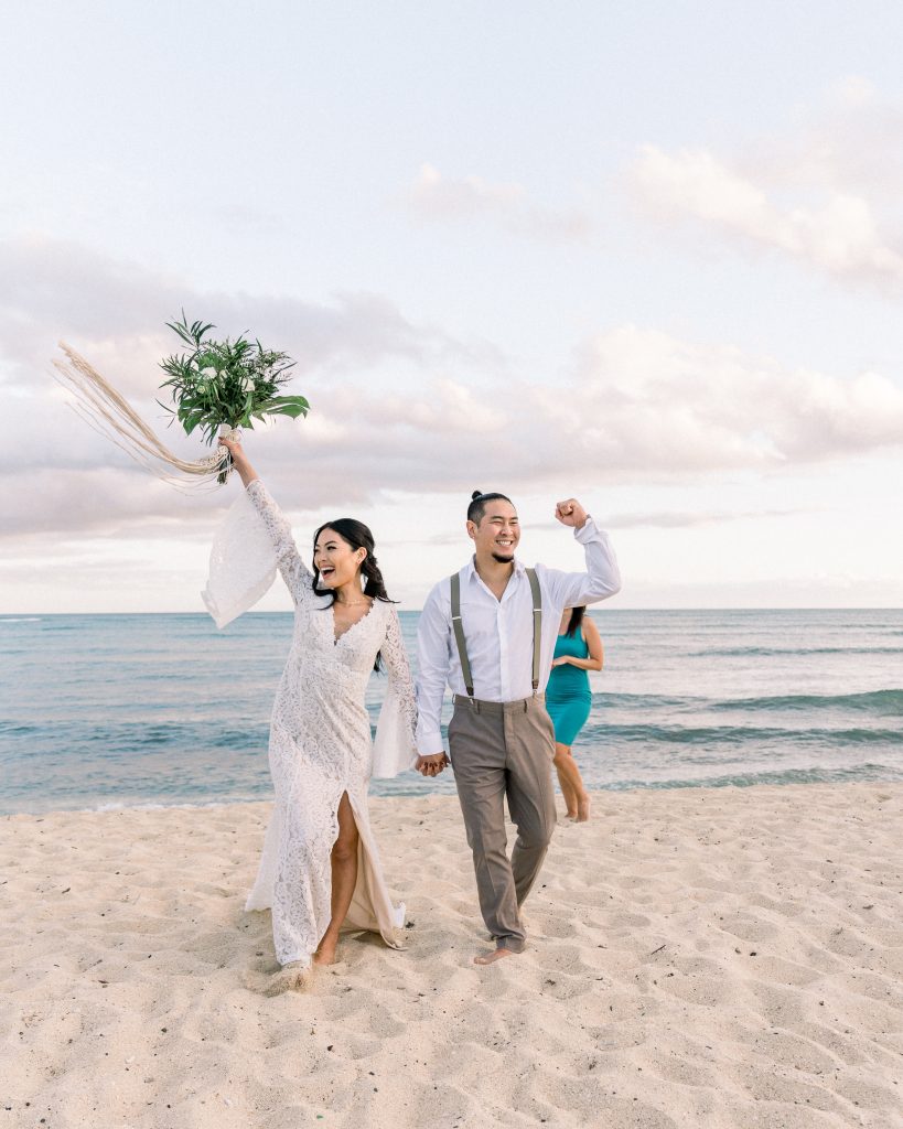 Bride and groom cheering after eloping on the beach in Hawaii. Bride is holding bouquet above her head as she cheers and walks on the sand. Hawaii Beach Weddings elopement packages Oahu Hawaii by Melissa Meyer